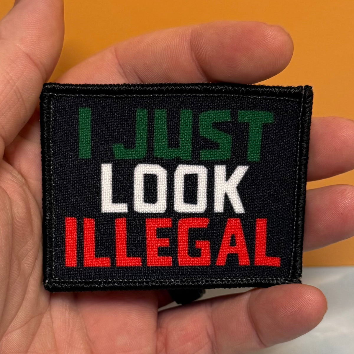 I Just Look Illegal printed canvas morale patch held in a hand, featuring the design lettering in green, white, and red lettering on a black background.