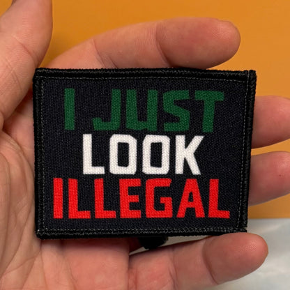 I Just Look Illegal printed canvas morale patch held in a hand, featuring the design lettering in green, white, and red lettering on a black background.