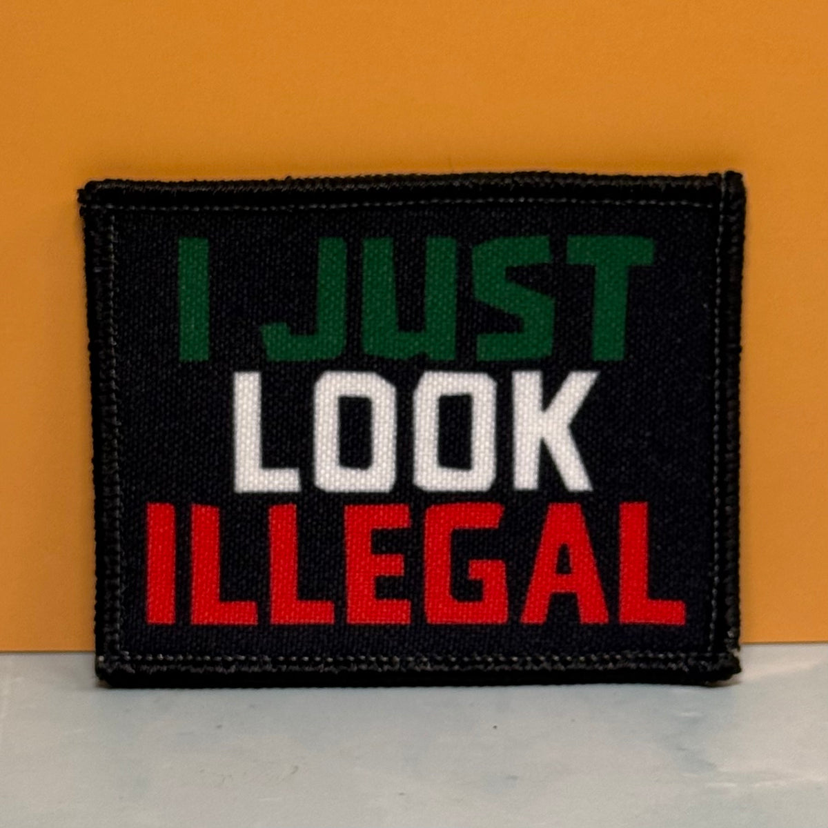 I Just Look Illegal printed canvas morale patch, design lettering in green, white, and red lettering, shown against an orange background.