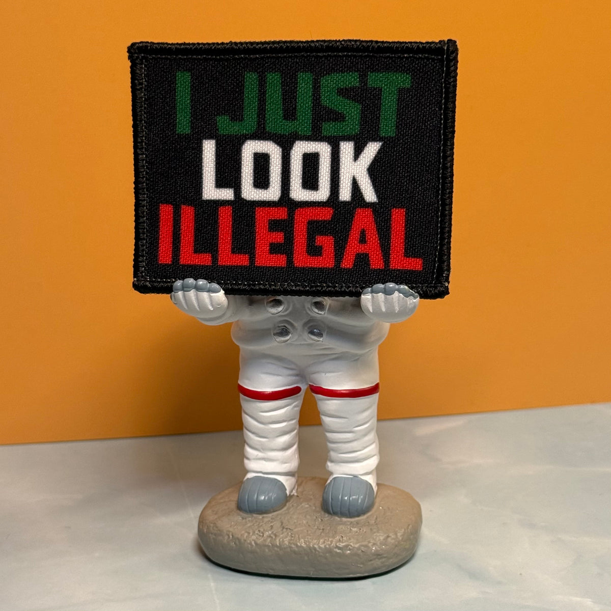 I Just Look Illegal printed canvas morale patch, a novelty figurine of a person in a white spacesuit holding a black rectangular sign that the design lettering in green, white, and red text, set against an orange background.