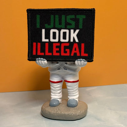 I Just Look Illegal printed canvas morale patch, a novelty figurine of a person in a white spacesuit holding a black rectangular sign that the design lettering in green, white, and red text, set against an orange background.