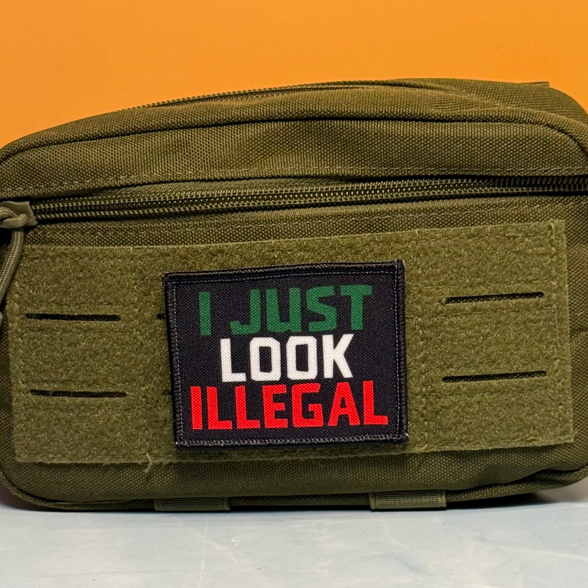 I Just Look Illegal printed canvas morale patch, an olive green tactical pouch with a front patch that the design lettering in green, white, and red text on a black background, set against an orange backdrop.