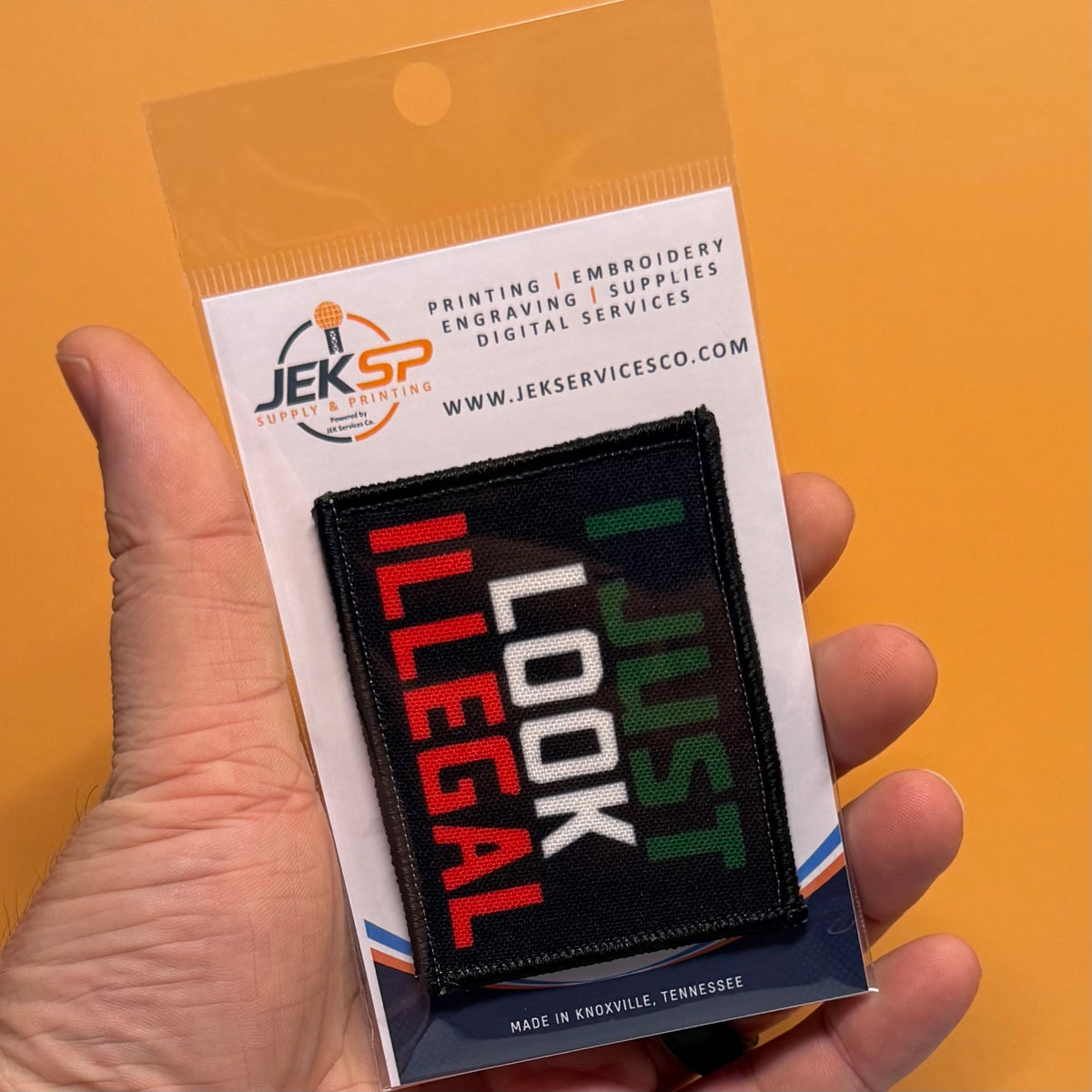 I Just Look Illegal printed canvas morale patch, a hand holds a packaged printed patch that the design lettering in red, white, and green text on a black background. The packaging card shows the Jeksp brand and includes text.