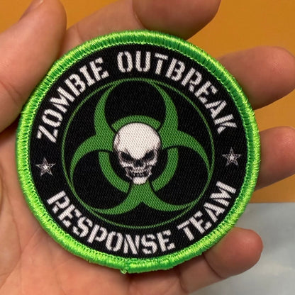 Zombie Outbreak Response Team printed canvas morale patch held in a hand, featuring a black background with a bright green border, a green biohazard symbol, a white skull in the center, and white text the design lettering.