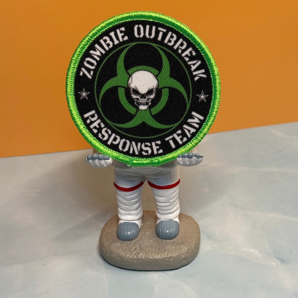 Zombie Outbreak Response Team printed canvas morale patch, the design lettering with a green biohazard symbol and skull in the center, held by a small astronaut figurine against an orange and gray background.