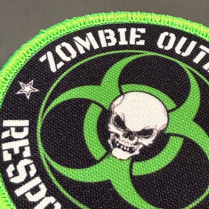 Zombie Outbreak Response Team printed canvas morale patch, a close-up of a circular printed patch featuring a black background, bright neon green border and biohazard-style symbol, a white skull in the center, and white text.