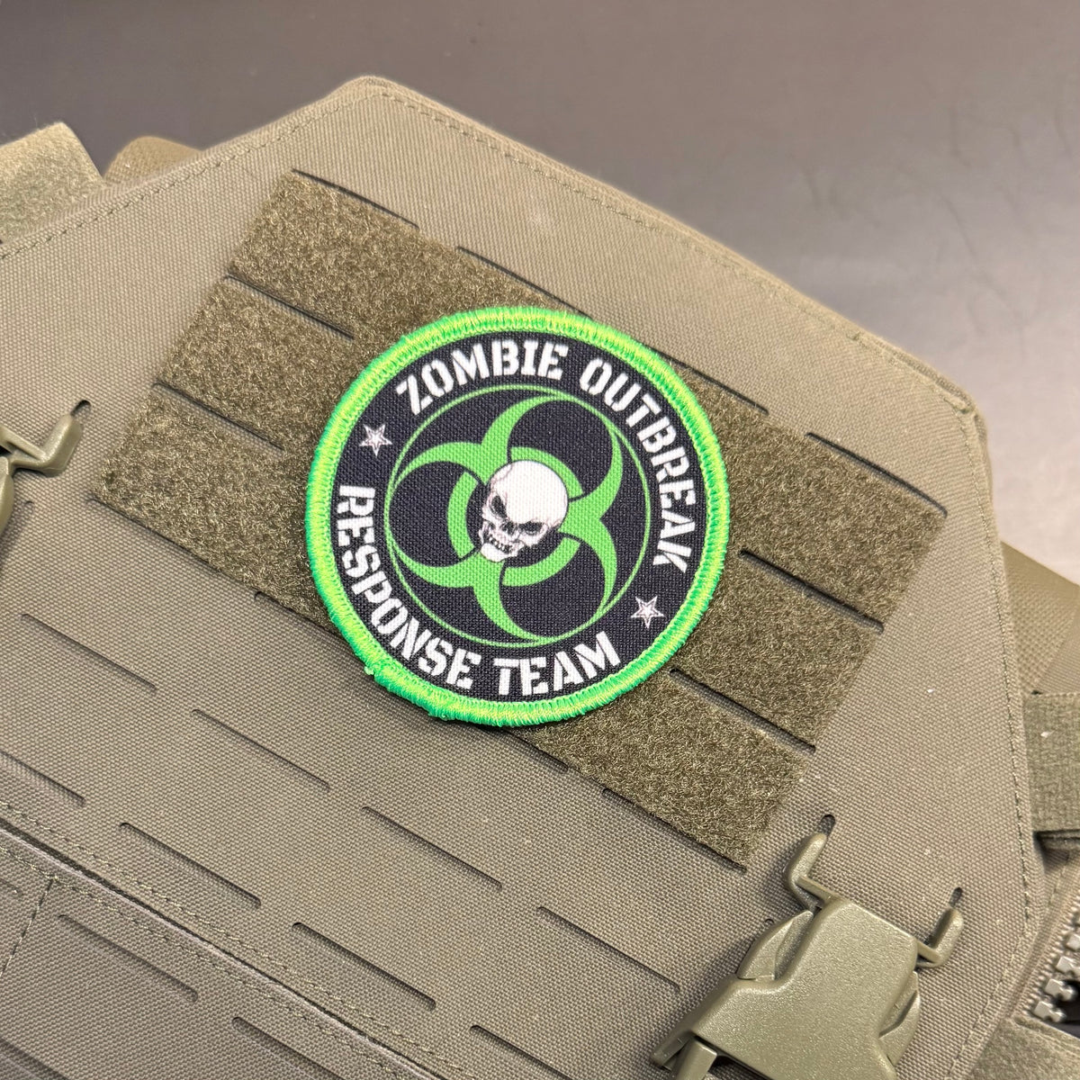 Zombie Outbreak Response Team printed canvas morale patch, the design lettering with a skull and biohazard symbol, attached to tan MOLLE webbing on a tan tactical vest or bag.