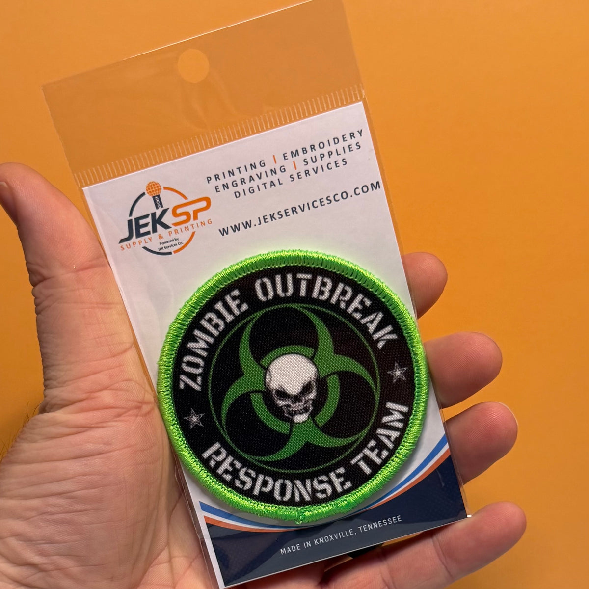Zombie Outbreak Response Team printed canvas morale patch, a hand holds a packaged printed patch against a solid orange background. The patch the design lettering with a skull and biohazard symbol in green, black, and white.