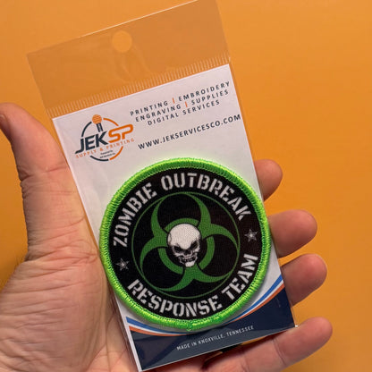 Zombie Outbreak Response Team printed canvas morale patch, a hand holds a packaged printed patch against a solid orange background. The patch the design lettering with a skull and biohazard symbol in green, black, and white.