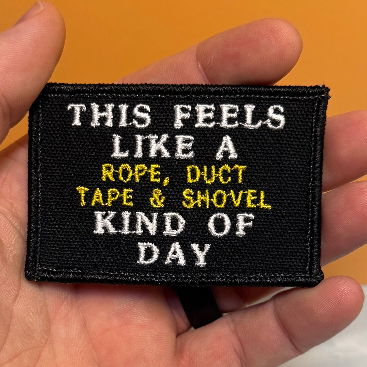This Feels Like A Rope & Duct Tape Kind Of Day embroidered morale patch: held in a hand, featuring white and yellow text that reads, 'THIS FEELS LIKE A ROPE, DUCT TAPE & SHOVEL KIND OF DAY' against a warm.