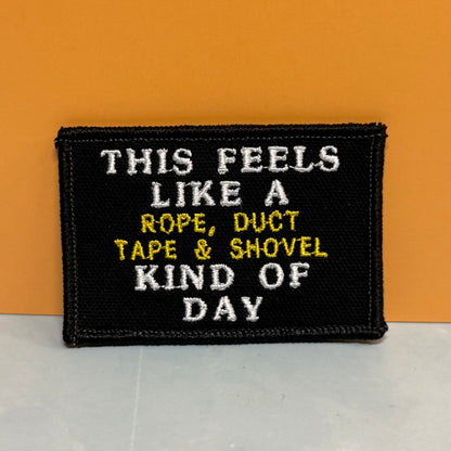 This Feels Like A Rope & Duct Tape Kind Of Day embroidered morale patch: black embroidered patch with white and yellow text reading 'THIS FEELS LIKE A ROPE, DUCT TAPE & SHOVEL KIND OF DAY', photographed against an orange.