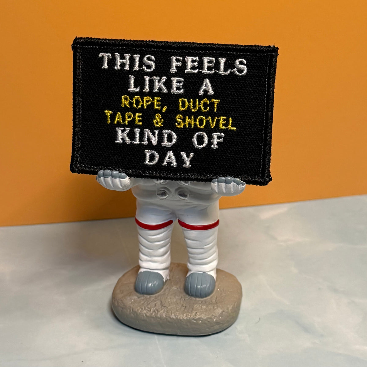 This Feels Like A Rope & Duct Tape Kind Of Day embroidered morale patch: novelty astronaut figurine holding a black embroidered patch with the text 'THIS FEELS LIKE A ROPE, DUCT TAPE & SHOVEL KIND OF DAY' in white and yellow.