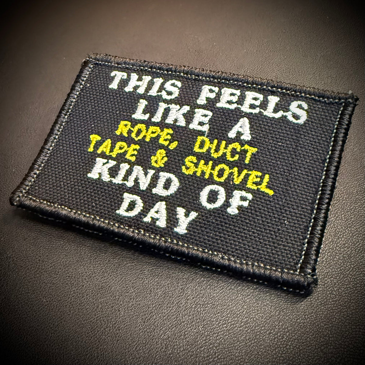 A rectangular embroidered patch with a black background and white and yellow text reading 'THIS FEELS LIKE A ROPE, DUCT TAPE & SHOVEL DAY' on a dark textured surface.