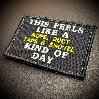 A rectangular embroidered patch with a black background and white and yellow text reading 'THIS FEELS LIKE A ROPE, DUCT TAPE & SHOVEL DAY' on a dark textured surface.