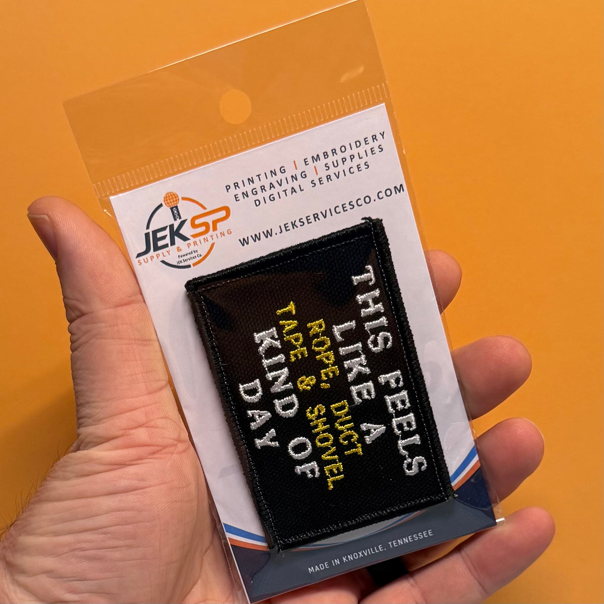 This Feels Like A Rope & Duct Tape Kind Of Day embroidered morale patch: hand holds a packaged black embroidered patch with the phrase “THIS FEELS LIKE A RIDE, DUCT TYPE & SHIELT KIND OF DAY” in white and yellow text, against a.