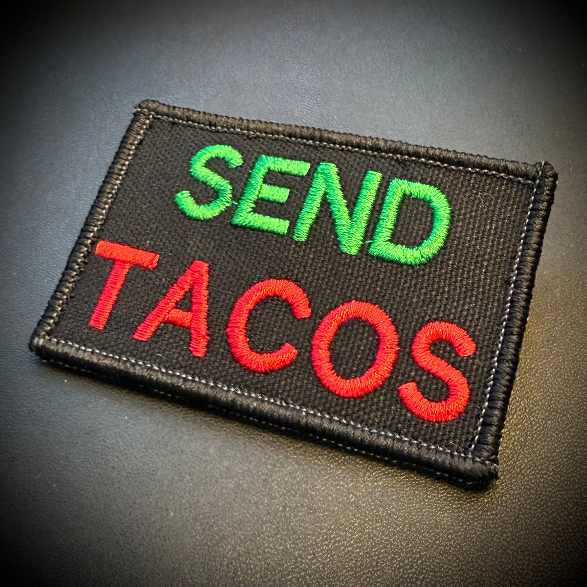 A rectangular embroidered patch with a black fabric border and the words 'SEND TACOS' in bright green and red lettering, photographed on a dark textured surface.