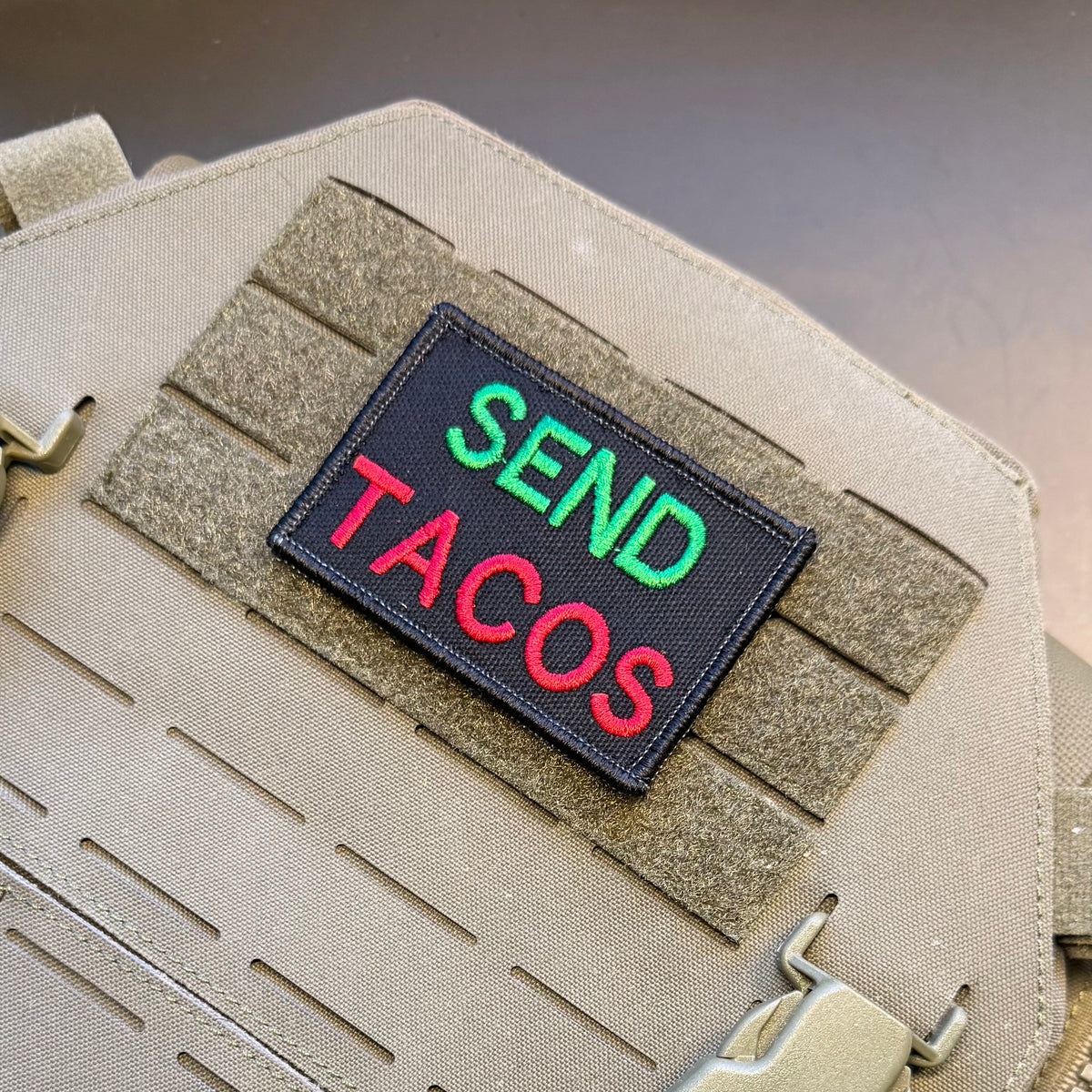 A rectangular embroidered patch reading 'SEND TACOS' in green and red lettering on a black background, attached to tan tactical webbing on a beige fabric item.