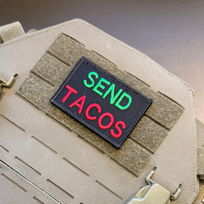A rectangular embroidered patch reading 'SEND TACOS' in green and red lettering on a black background, attached to tan tactical webbing on a beige fabric item.