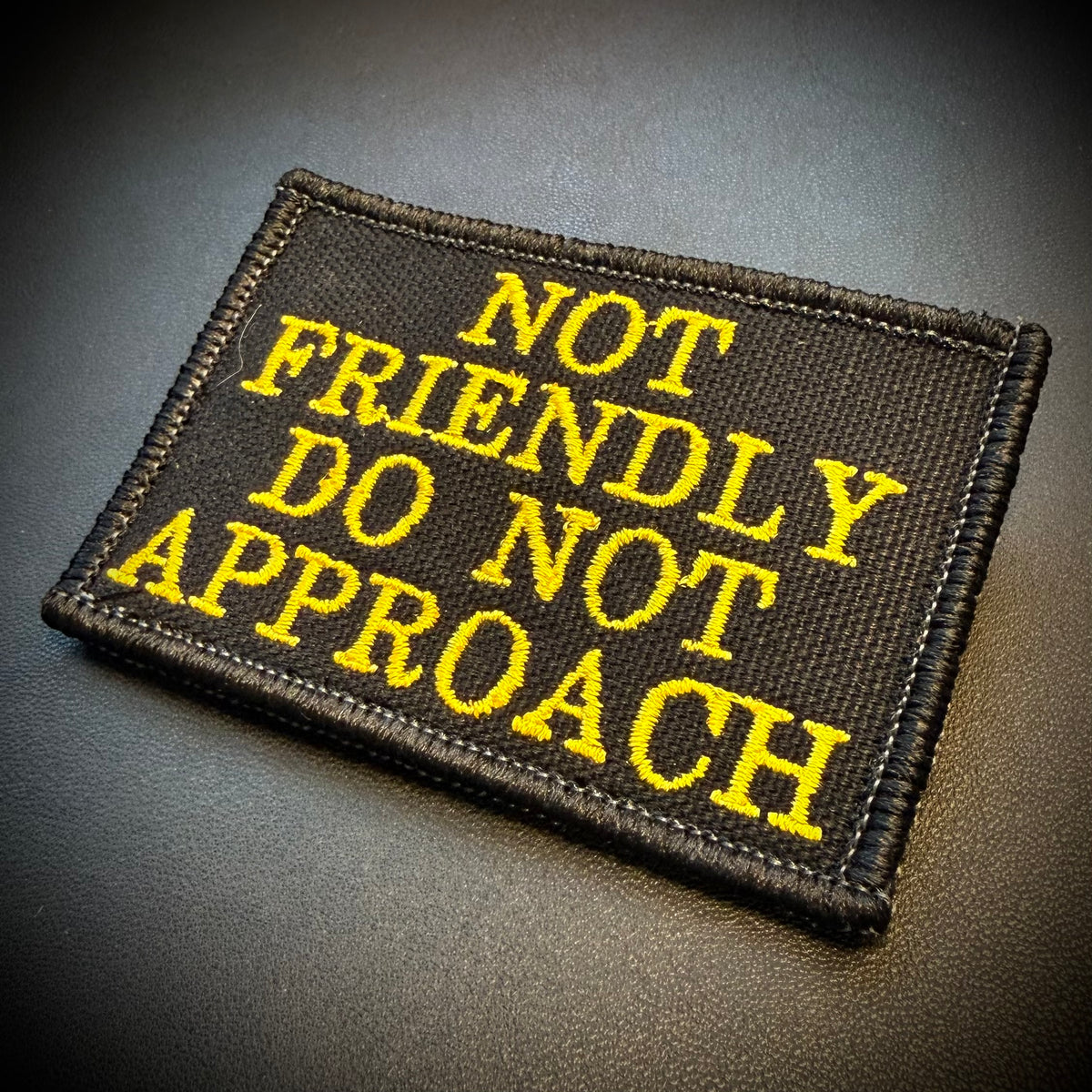 A rectangular black embroidered patch with yellow text reading 'NOT FRIENDLY DO NOT APPROACH' on a dark textured background.