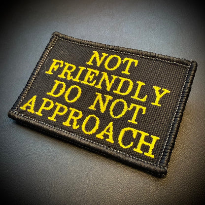 A rectangular black embroidered patch with yellow text reading 'NOT FRIENDLY DO NOT APPROACH' on a dark textured background.