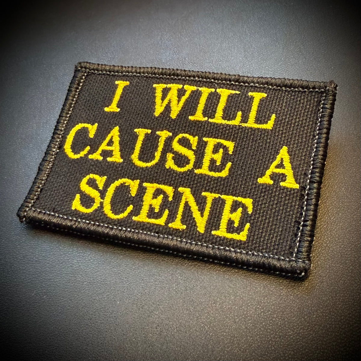 A rectangular embroidered patch with the text 'I WILL CAUSE A SCENE' in bright yellow lettering on a black fabric background, photographed on a dark textured surface.