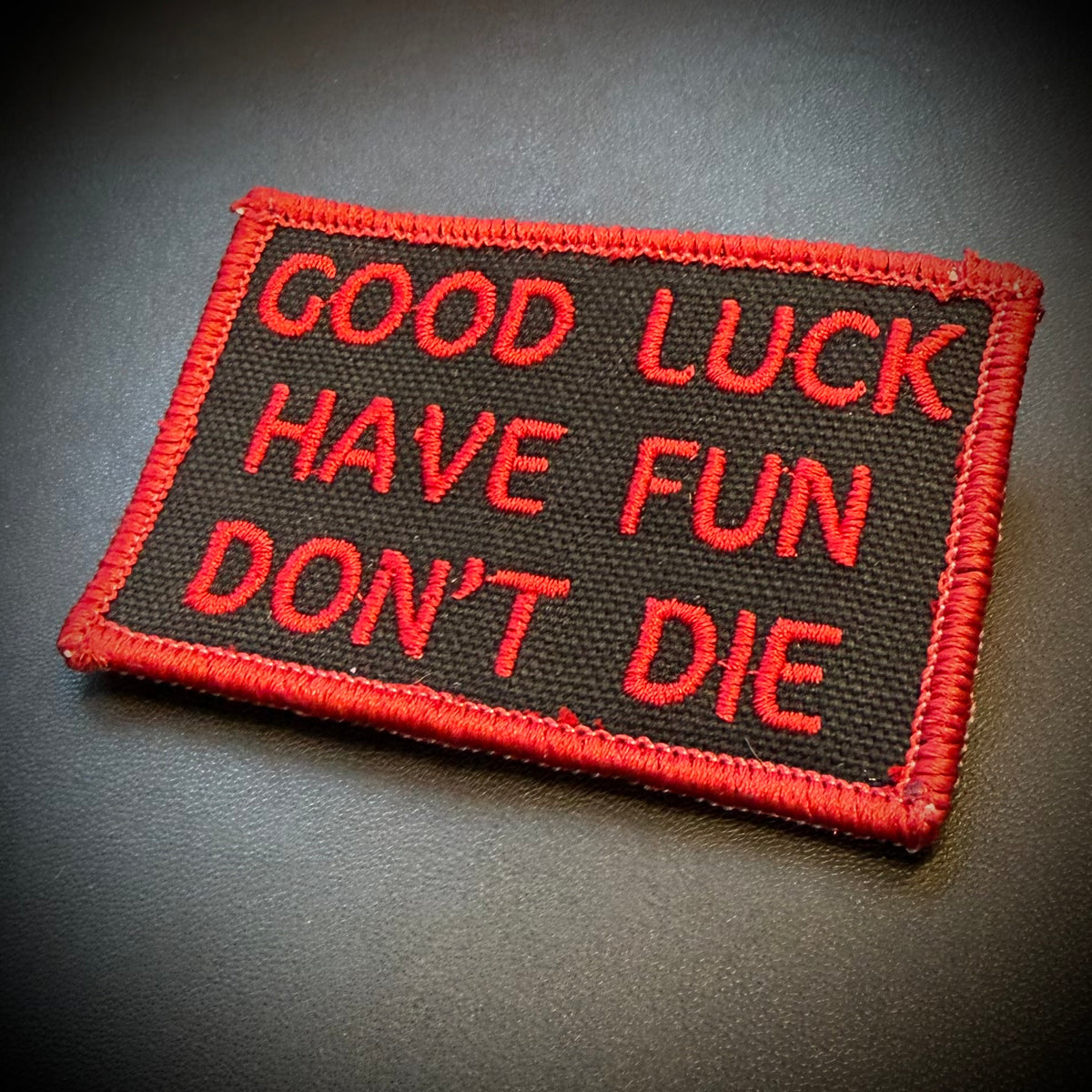 A rectangular embroidered patch with a black background and red border, featuring the text 'GOOD LUCK HAVE FUN DON'T DIE' in red embroidery, shown on a dark gray surface.