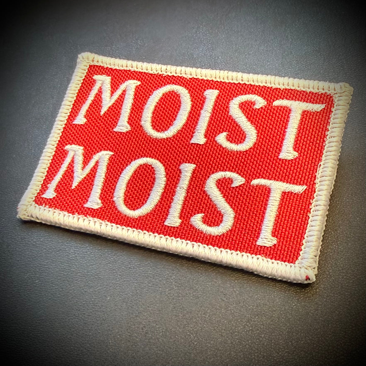 A rectangular embroidered patch with a red background and cream lettering that reads 'MOIST MOIST', finished with a cream stitched border, shown on a dark gray textured surface.