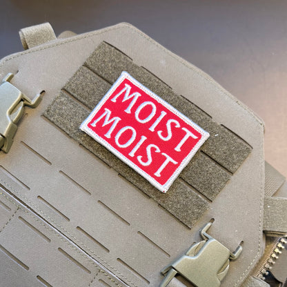 A rectangular embroidered patch with the words 'MOIST MOIST' in white on a red background, attached to a tan tactical vest with MOLLE webbing and quick-release buckles.