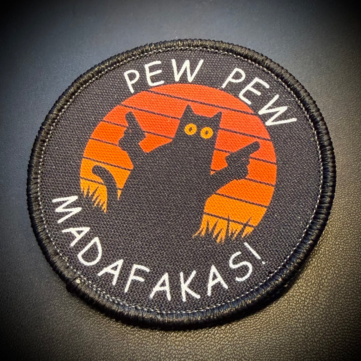 A circular embroidered patch with a black cat silhouette in front of an orange sunset, with the text 'PEW PEW MADAFAKASI' in white around the border.