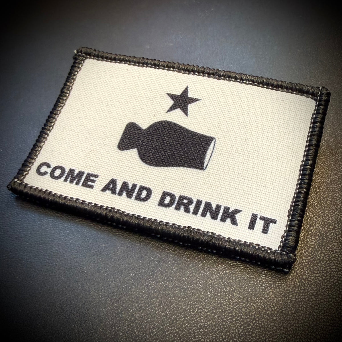 A rectangular embroidered patch with a black stitched border, featuring a black bottle graphic, a black star, and the text 'COME AND DRINK IT' on an off-white background, placed on a dark textured surface.
