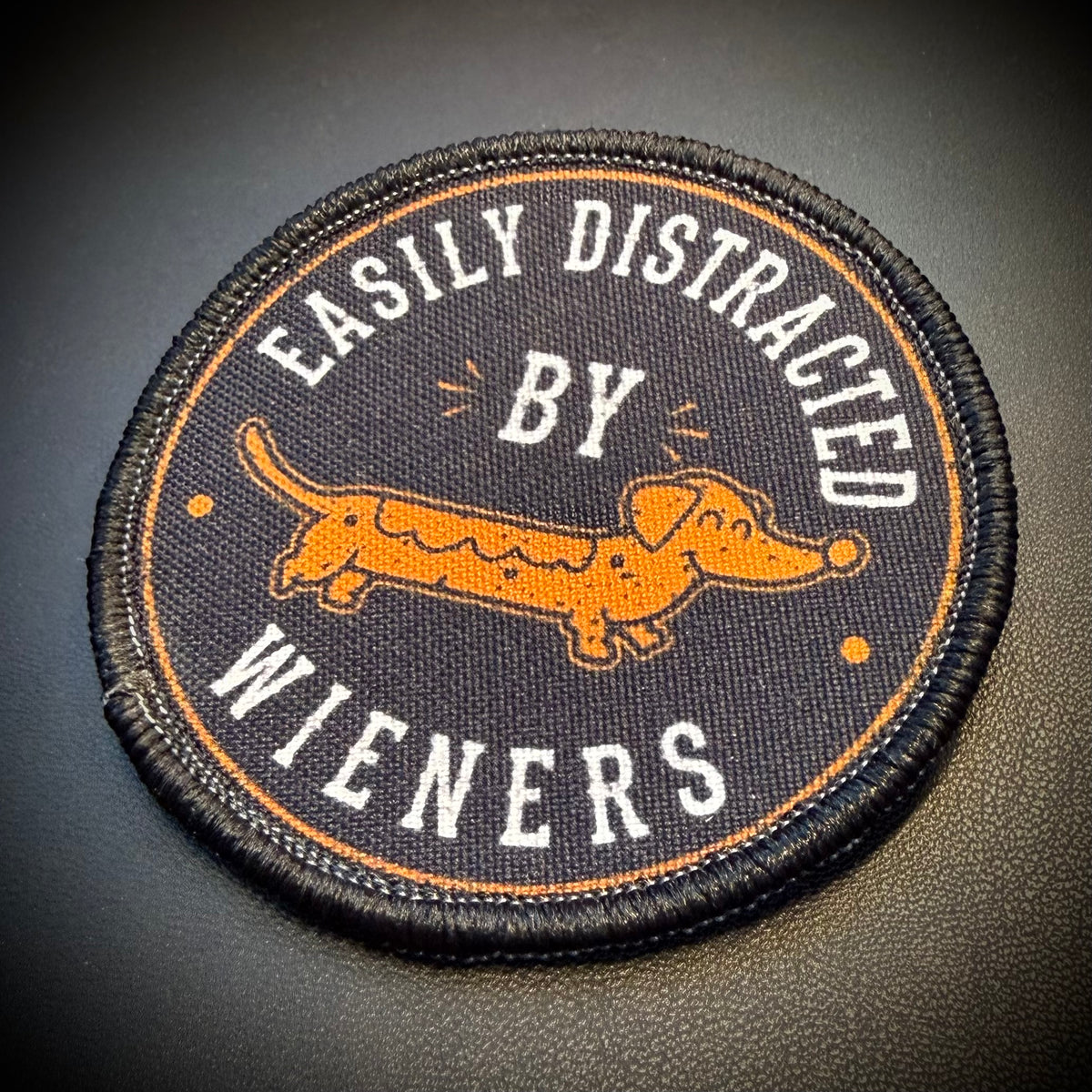 A circular embroidered patch featuring a dachshund illustration and the text 'EASILY DISTRACTED BY WIENERS' in white on a dark background, photographed on a dark textured surface.