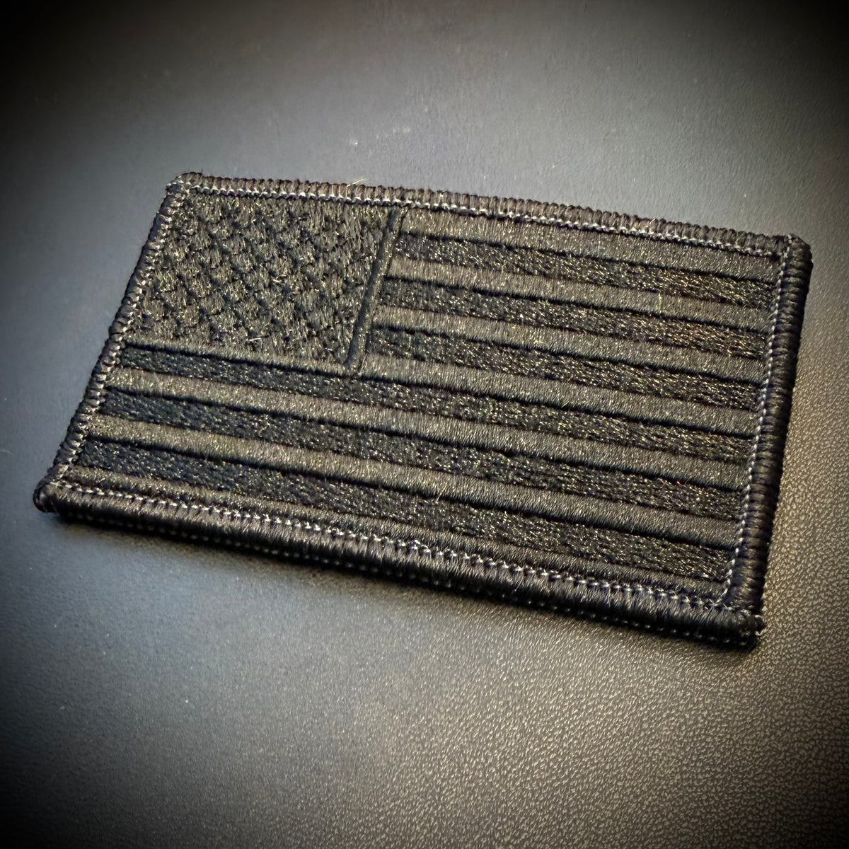 A rectangular embroidered American flag patch in black and dark gray, shown on a dark textured surface.
