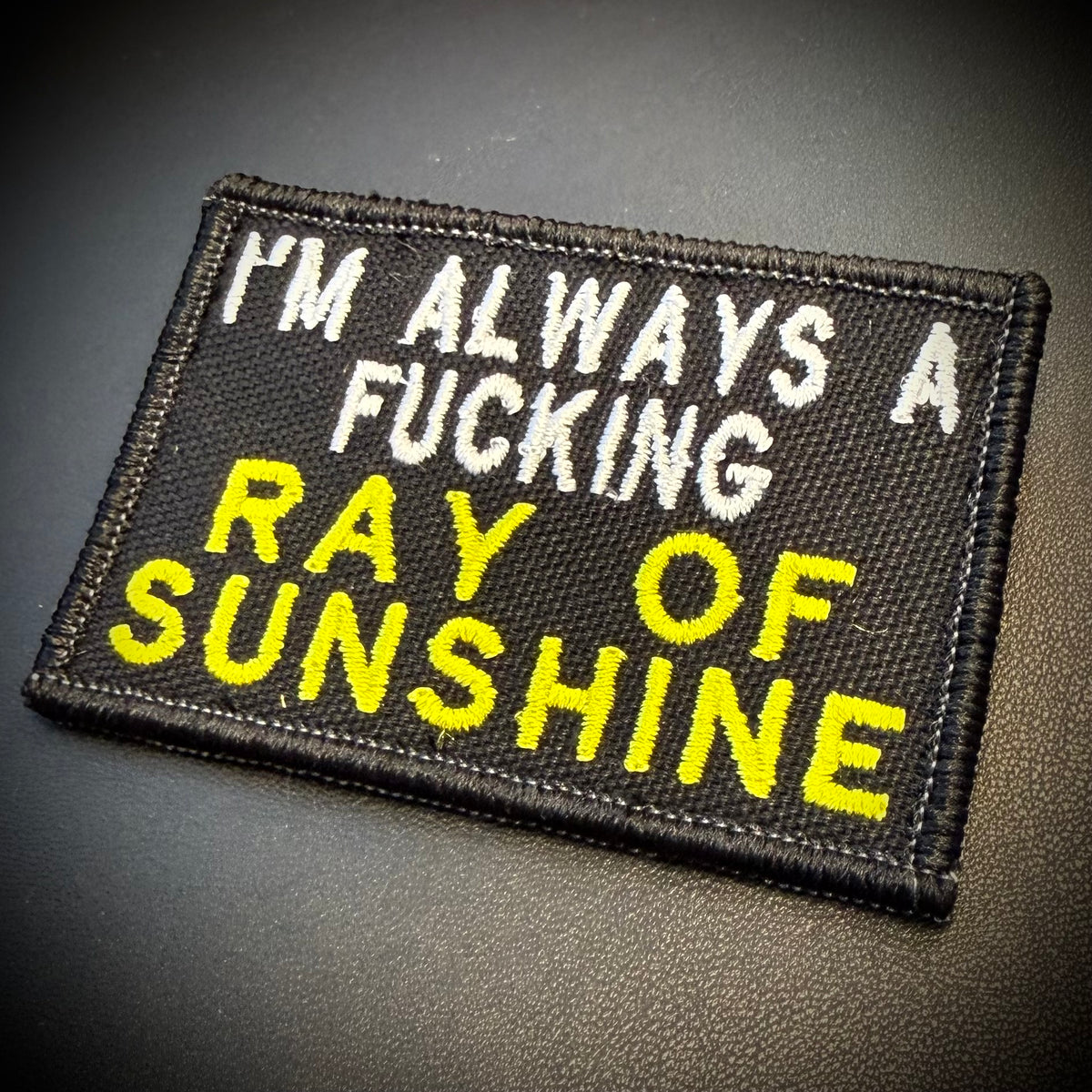 A rectangular embroidered patch with black background, white text reading 'I'M ALWAYS A', and large yellow text reading 'RAY OF SUNSHINE' on a dark textured surface.