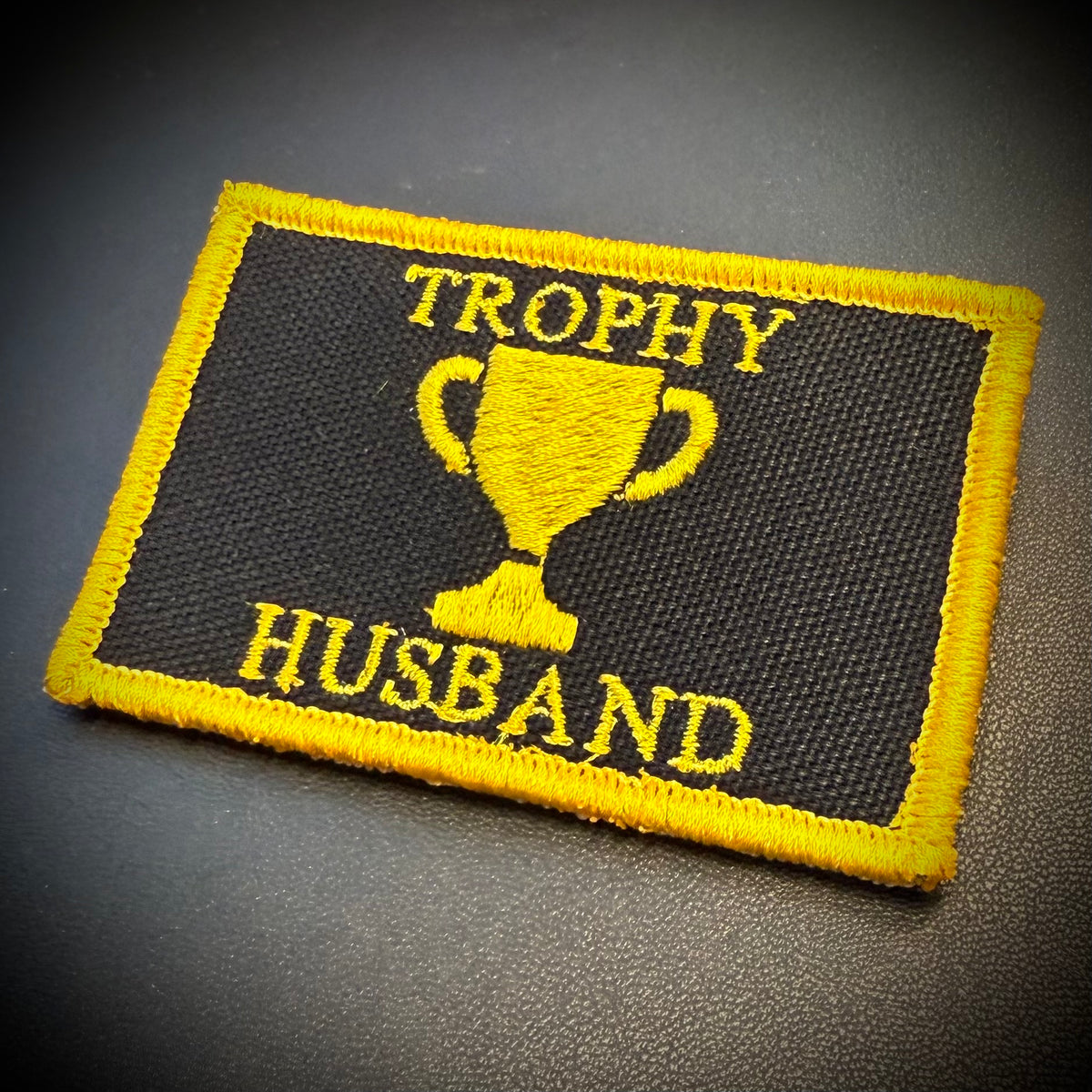 A rectangular embroidered patch with a black background and yellow border, featuring a yellow trophy graphic and the words 'TROPHY HUSBAND' in yellow thread, photographed on a dark textured surface.