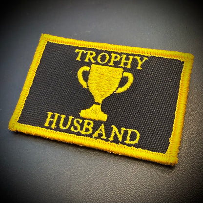 A rectangular embroidered patch with a black background and yellow border, featuring a yellow trophy graphic and the words 'TROPHY HUSBAND' in yellow thread, photographed on a dark textured surface.