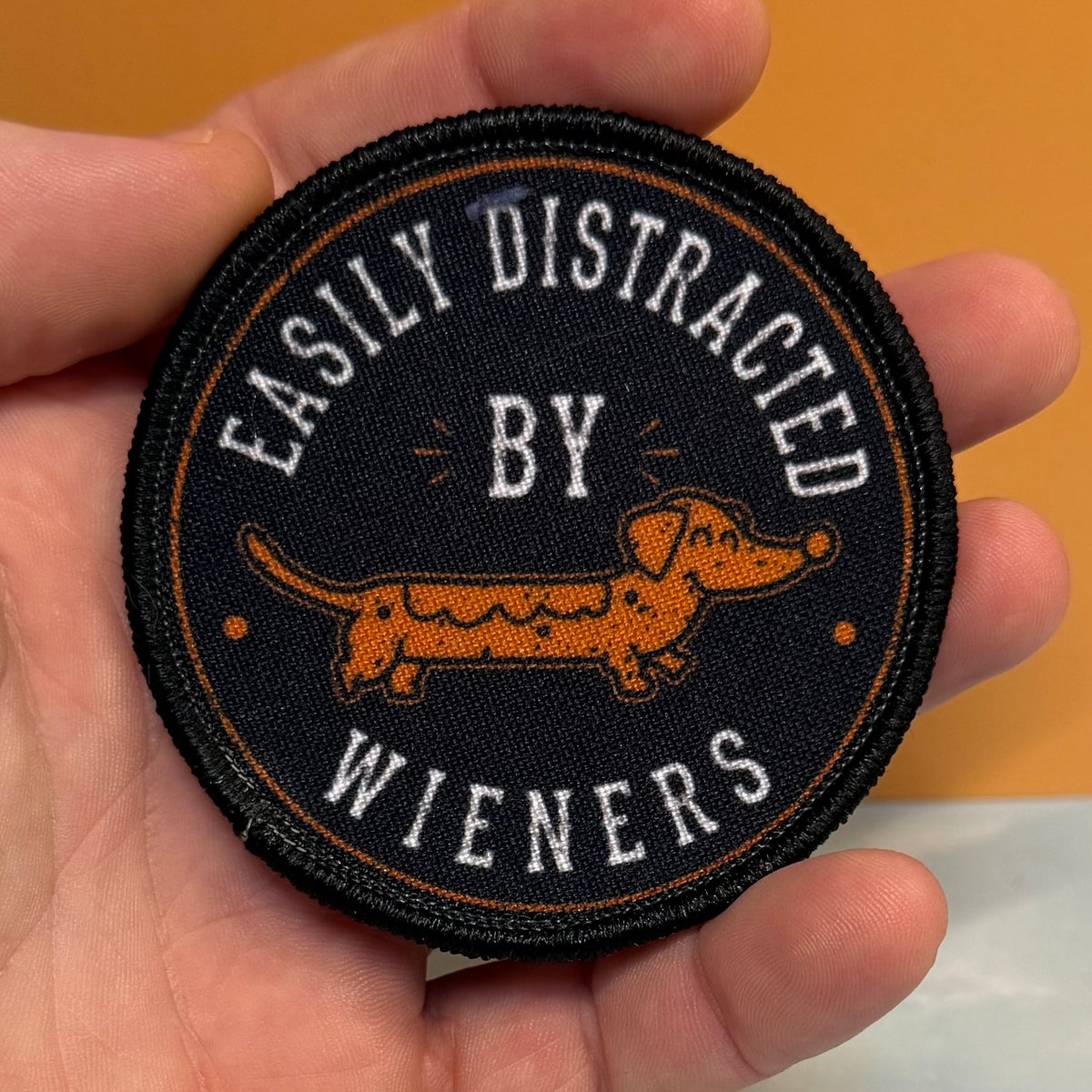 A round embroidered patch held in a hand, featuring a black background with a brown dachshund and white text reading 'EASILY DISTRACTED BY WIENERS'.