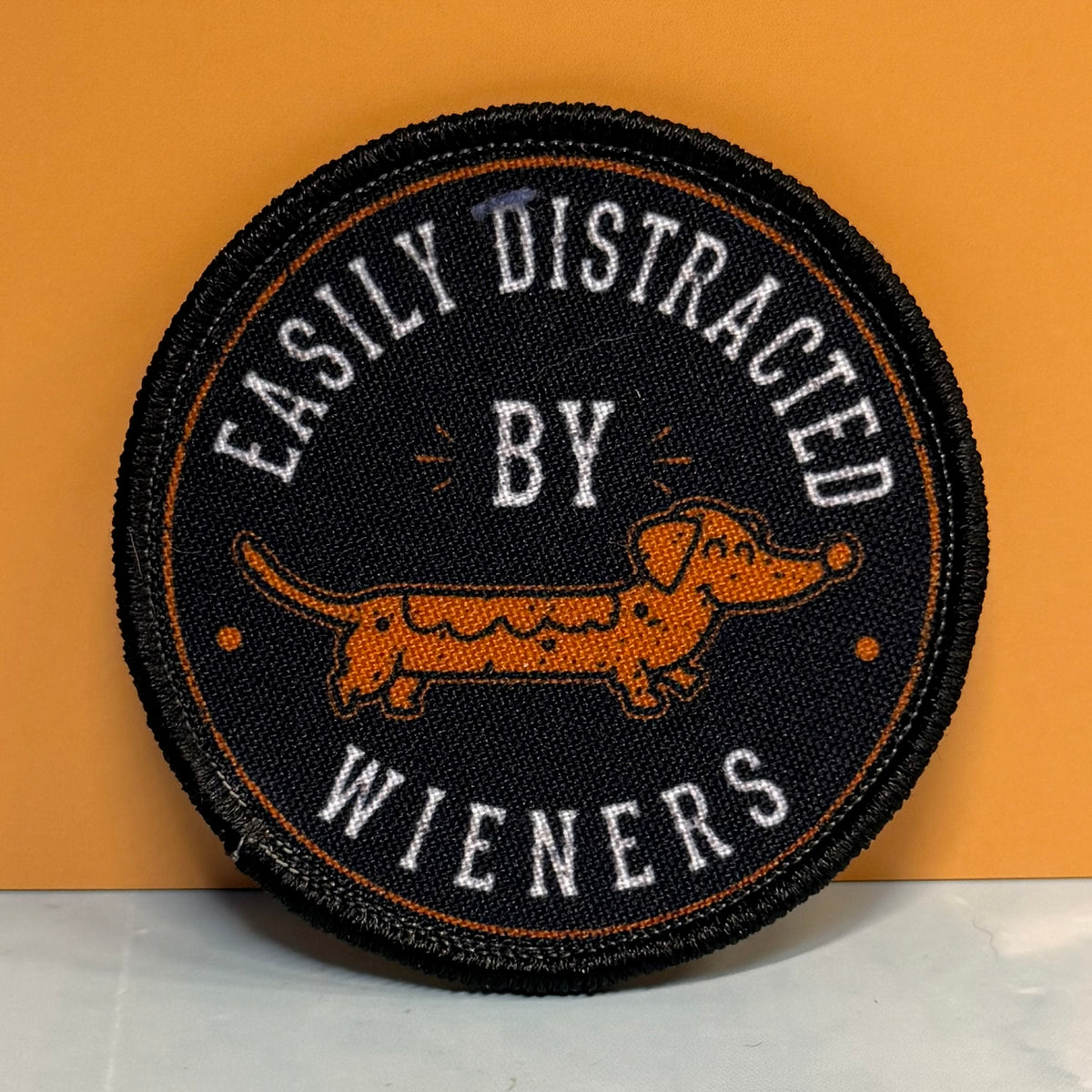 A round embroidered patch with a black background, featuring a dachshund in orange and white text that reads 'EASILY DISTRACTED BY WIENERS'.
