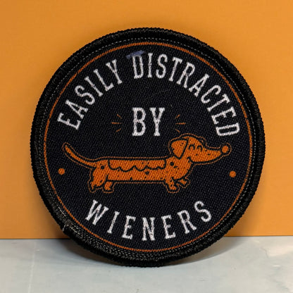 A round embroidered patch with a black background, featuring a dachshund in orange and white text that reads 'EASILY DISTRACTED BY WIENERS'.