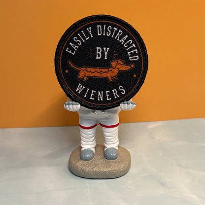 A novelty astronaut figurine holding a round black patch with a dachshund and the text 'EASILY DISTRACTED BY WIENERS', set against an orange and white background.
