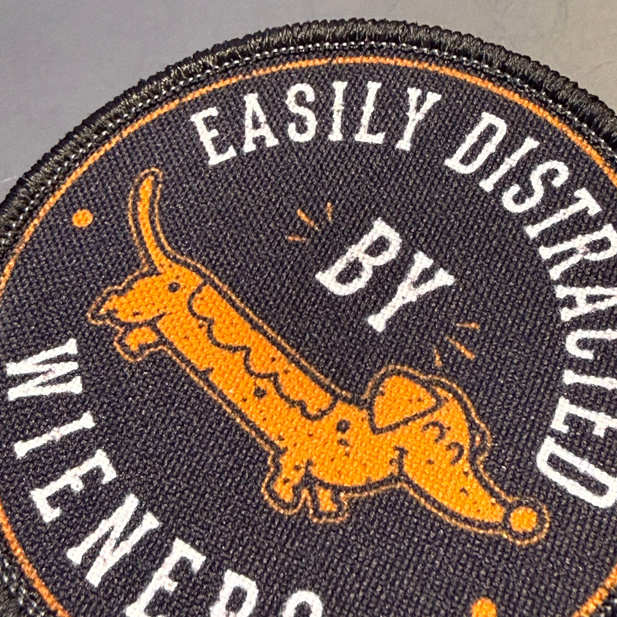 A close-up of a circular embroidered patch featuring a dachshund illustration and the text 'EASILY DISTRALED BY WIENERS' in white on a dark background.