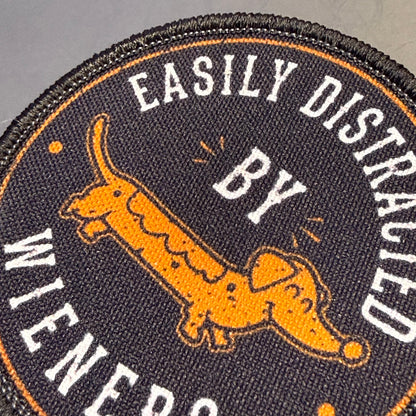 A close-up of a circular embroidered patch featuring a dachshund illustration and the text 'EASILY DISTRALED BY WIENERS' in white on a dark background.