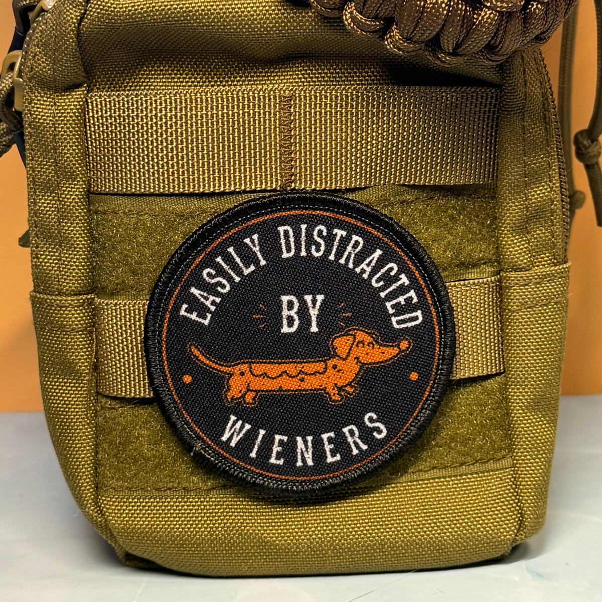 A close-up of a tactical pouch in olive drab green webbed fabric, featuring a circular embroidered patch with a dachshund and the text 'EASILY DISTRACTED BY WIENERS'.