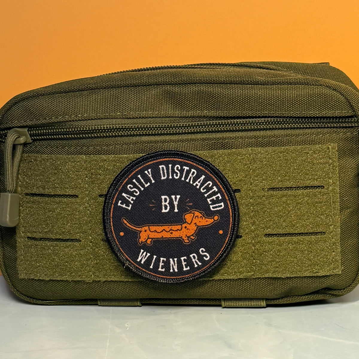 An olive green tactical pouch with a circular embroidered patch reading 'EASILY DISTRACTED BY WIENERS' and featuring an orange dachshund, set against a solid orange background.