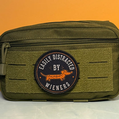 An olive green tactical pouch with a circular embroidered patch reading 'EASILY DISTRACTED BY WIENERS' and featuring an orange dachshund, set against a solid orange background.