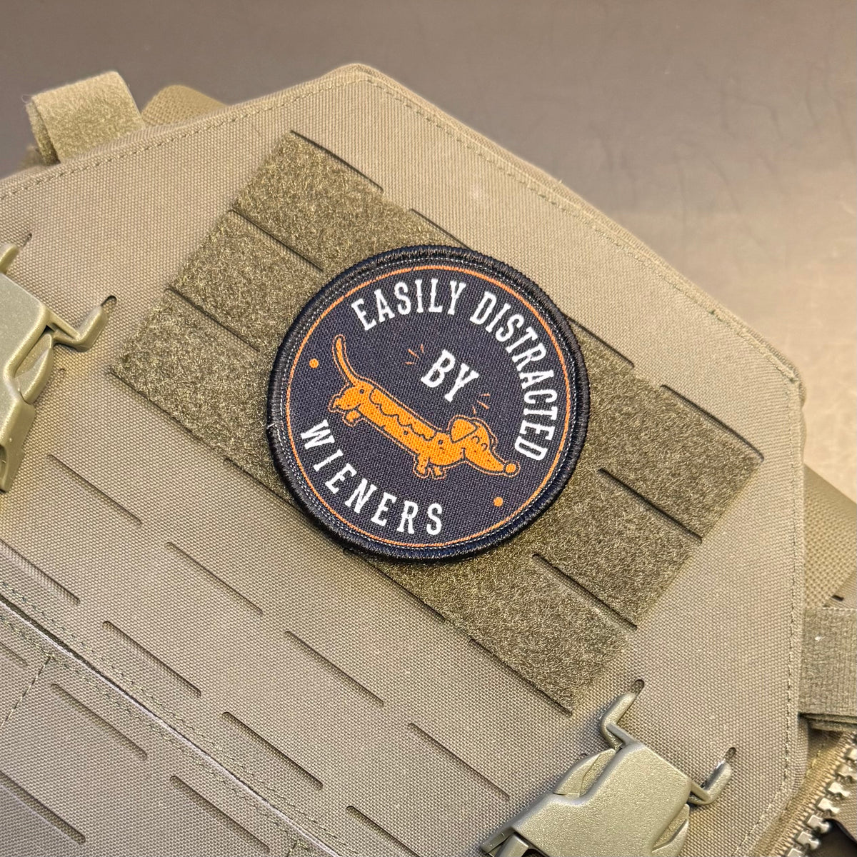 A circular embroidered patch with a dachshund and the text 'EASILY DISTRACED BY WIENERS' in orange and white on a dark navy background, attached to a tan tactical vest with Velcro strips and metal buckles.