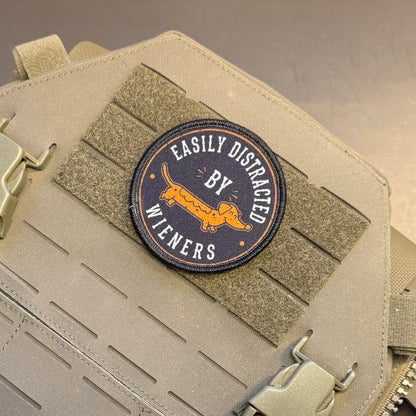 A circular embroidered patch with a dachshund and the text 'EASILY DISTRACED BY WIENERS' in orange and white on a dark navy background, attached to a tan tactical vest with Velcro strips and metal buckles.