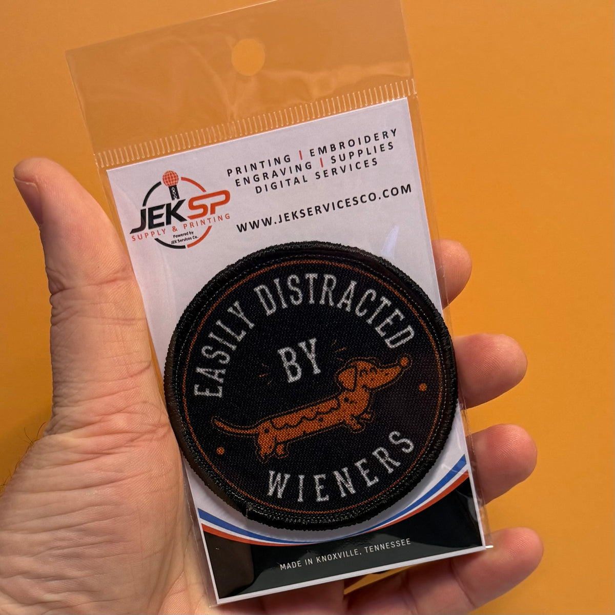 A hand holding a clear plastic blister pack containing an embroidered oval patch. The patch reads “EASILY DISTRACTED BY WIENERS” and shows a dachshund illustration.