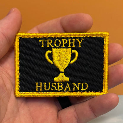 A rectangular embroidered patch held in a hand, featuring a black background with a yellow border, a yellow trophy graphic, and the words 'TROPHY HUSBAND' in yellow embroidery.