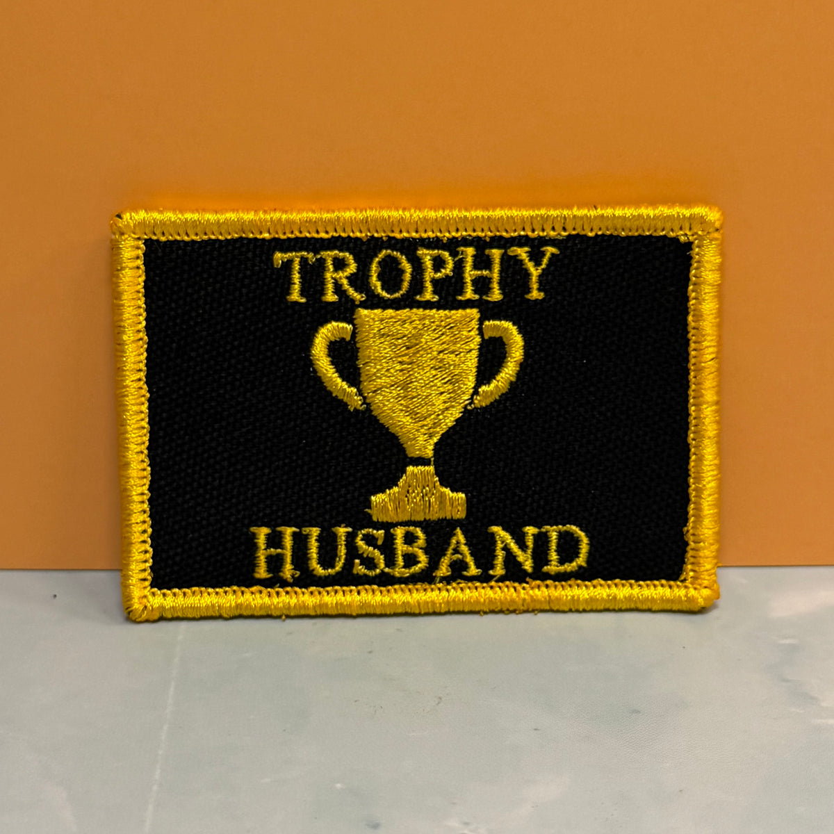 A rectangular embroidered patch with a black background, yellow border, and yellow text reading 'TROPHY HUSBAND' with a trophy graphic in the center, placed against an orange wall and light gray surface.