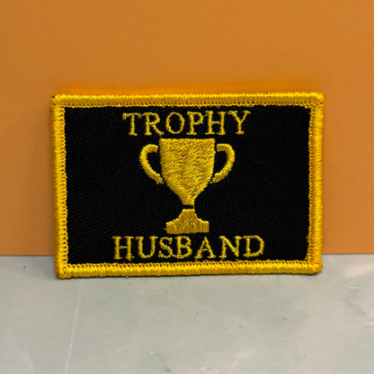 A rectangular embroidered patch with a black background, yellow border, and yellow text reading 'TROPHY HUSBAND' with a trophy graphic in the center, placed against an orange wall and light gray surface.